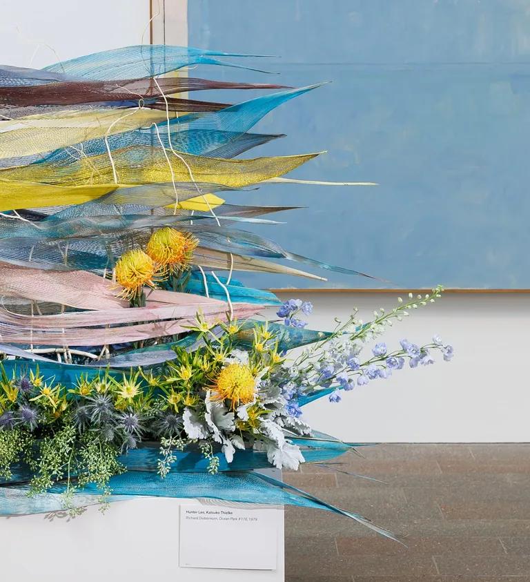 A floral arrangement on a woven blue and yellow sculpture in front of an abstract painting