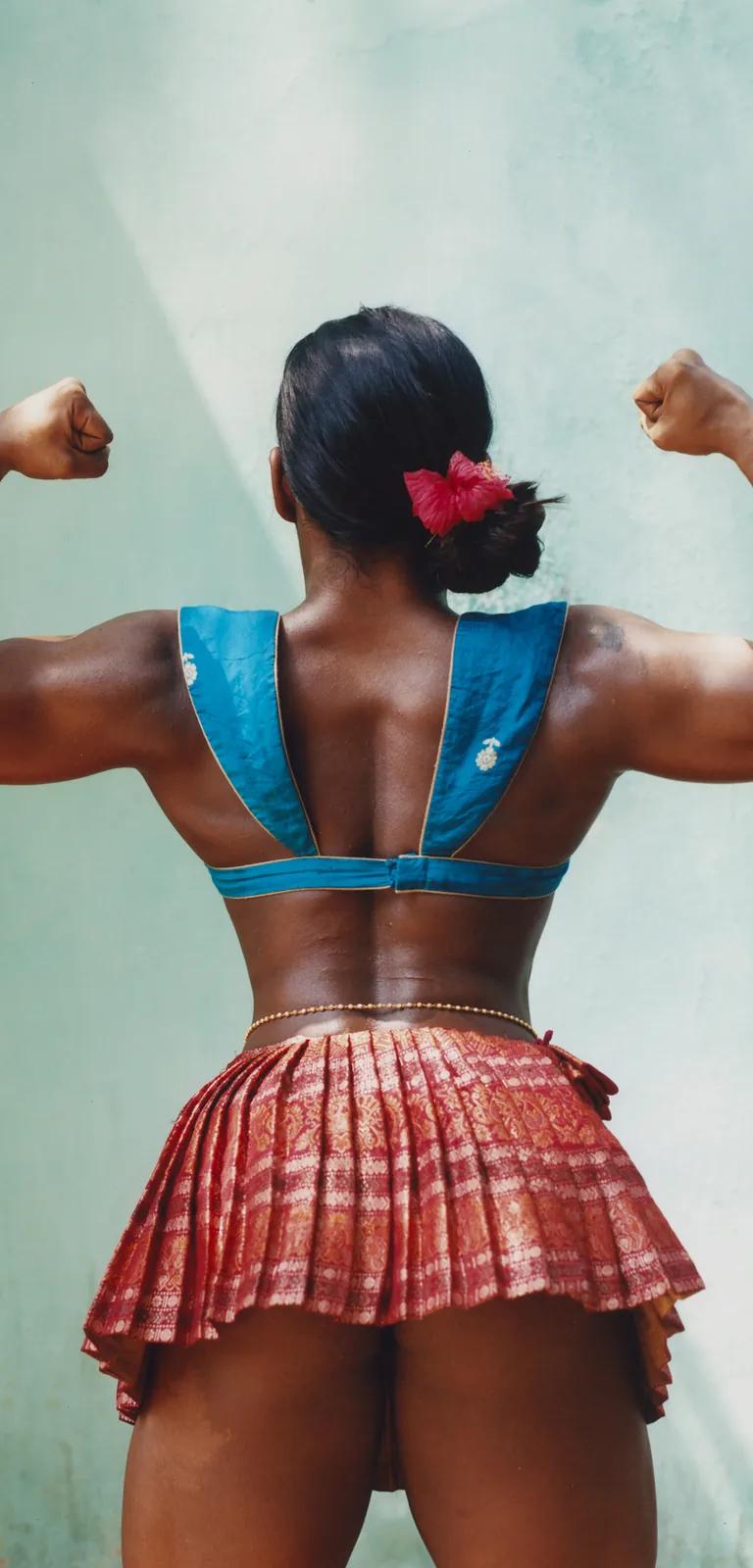 Bhumika, 2024: a woman in a sleeveless green dress flexing her arm outdoors among palm trees in Kerala, by Keerthana Kunnath