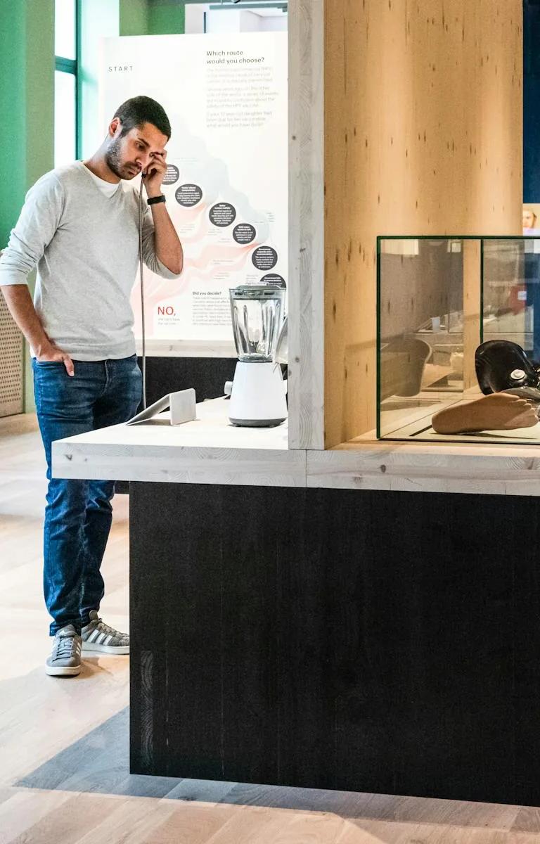 Photograph of an exhibition gallery space; a woman looks at objects in a glass case while touching an info panel, and a man holds a headphone to his ear listening to audio.