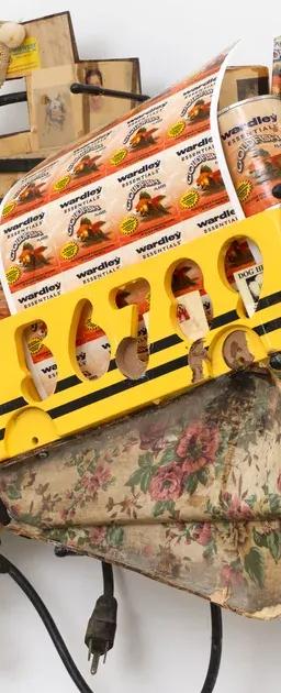 A wall-mounted assemblage sculpture of yellow toy buses, black electrical cords, white beads and small photographs on a floral-patterned fabric base