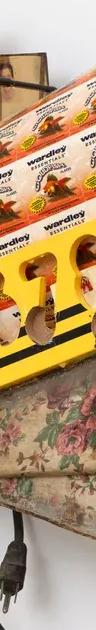 A wall-mounted assemblage sculpture of yellow toy buses, black electrical cords, white beads and small photographs on a floral-patterned fabric base