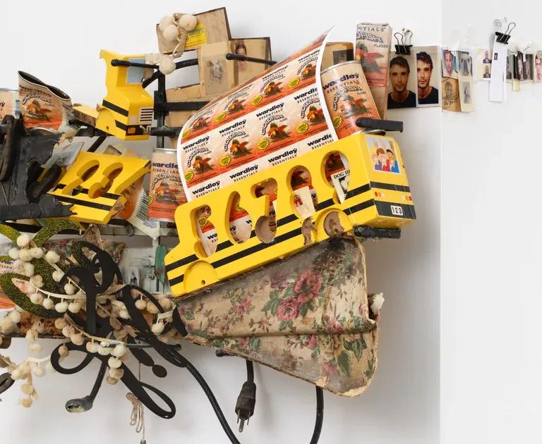 A wall-mounted assemblage sculpture of yellow toy buses, black electrical cords, white beads and small photographs on a floral-patterned fabric base