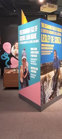 Museum exhibitions featuring two scientists. On the left is a desk and bookshelves, with multimedia panels. On the right, a large video screen and a canoe.