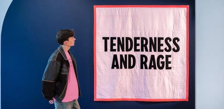 A visitor at the exhibition entrance looking at a large light pink quilt with a bright red hem, with the words 'Tenderness and Rage' sewn onto it in capital letters.