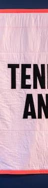 A visitor at the exhibition entrance looking at a large light pink quilt with a bright red hem, with the words 'Tenderness and Rage' sewn onto it in capital letters.
