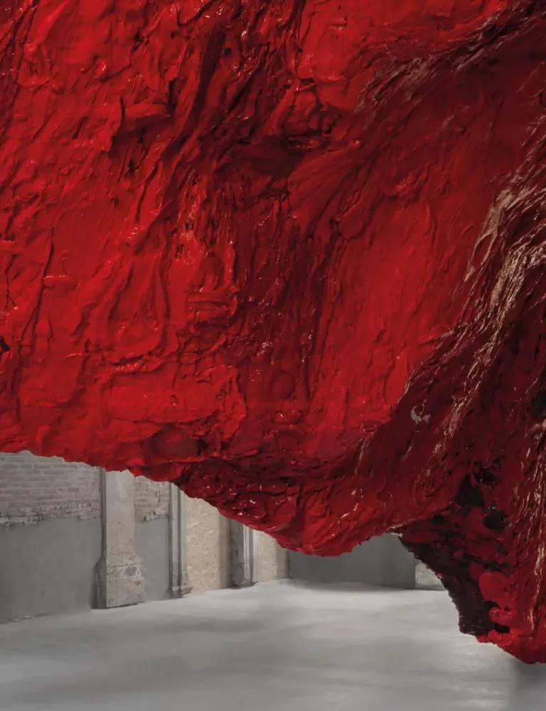 Large red textured sculpture by Anish Kapoor hanging from ceiling to floor in a columned space