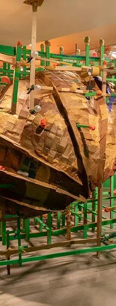 A large, textured, brown cardboard sculpture encased within a green wooden grid structure, in a room with bright green walls and an orange t-shirt hanging from the ceiling