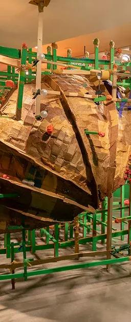 A large, textured, brown cardboard sculpture encased within a green wooden grid structure, in a room with bright green walls and an orange t-shirt hanging from the ceiling