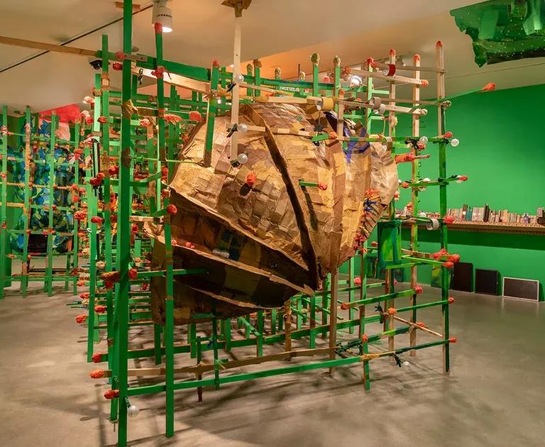 A large, textured, brown cardboard sculpture encased within a green wooden grid structure, in a room with bright green walls and an orange t-shirt hanging from the ceiling