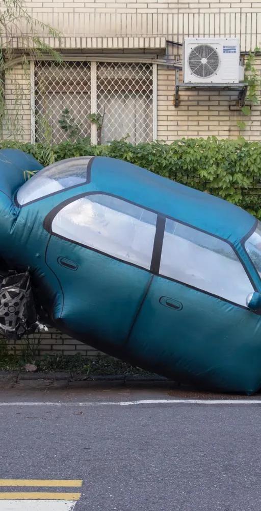 A man seemingly lifting the rear end of a large teal inflatable replica car wedged onto a curbside parking space in front of a residential building