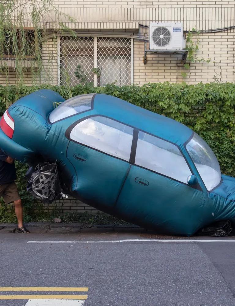 A man seemingly lifting the rear end of a large teal inflatable replica car wedged onto a curbside parking space in front of a residential building