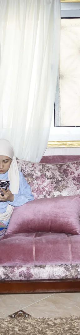 A woman on a pink floral sofa looking at her phone, from Yomna El Beyaly's series In Her Own Way