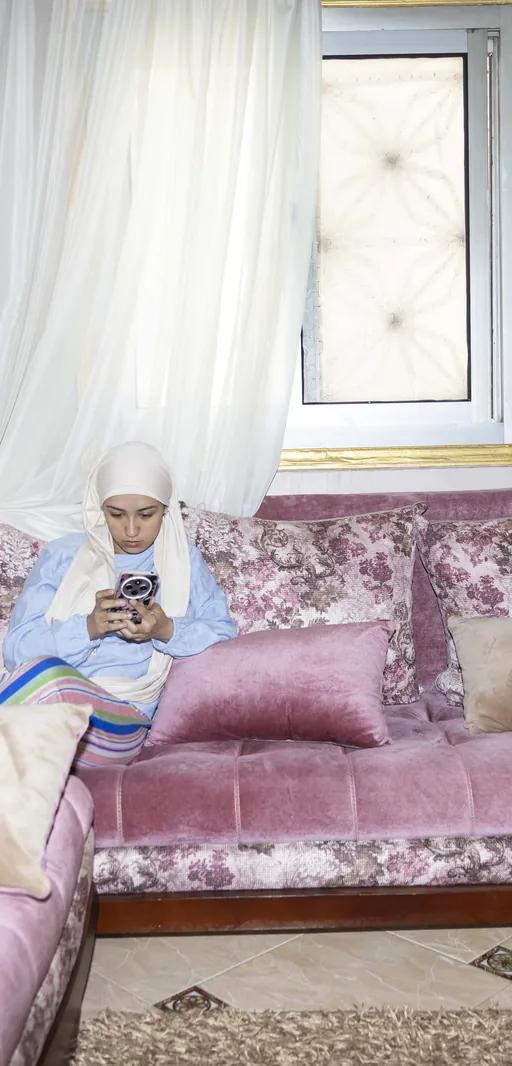 A woman on a pink floral sofa looking at her phone, from Yomna El Beyaly's series In Her Own Way