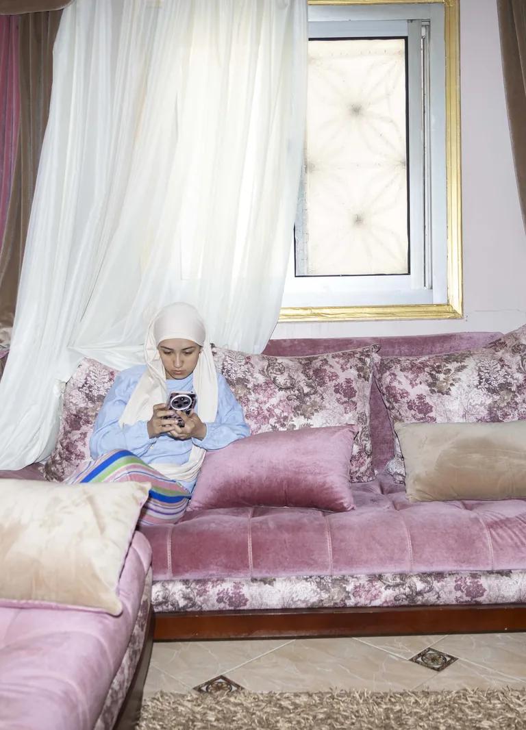 A woman on a pink floral sofa looking at her phone, from Yomna El Beyaly's series In Her Own Way