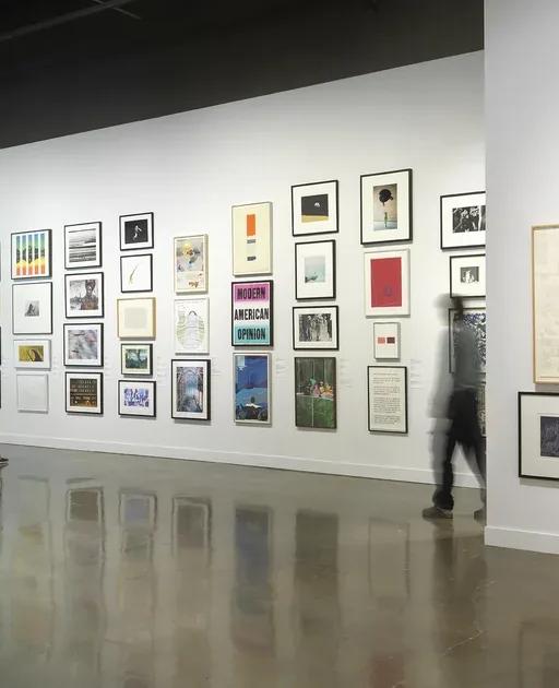 Installation view with framed artworks hung salon-style on the walls and three people looking at the art