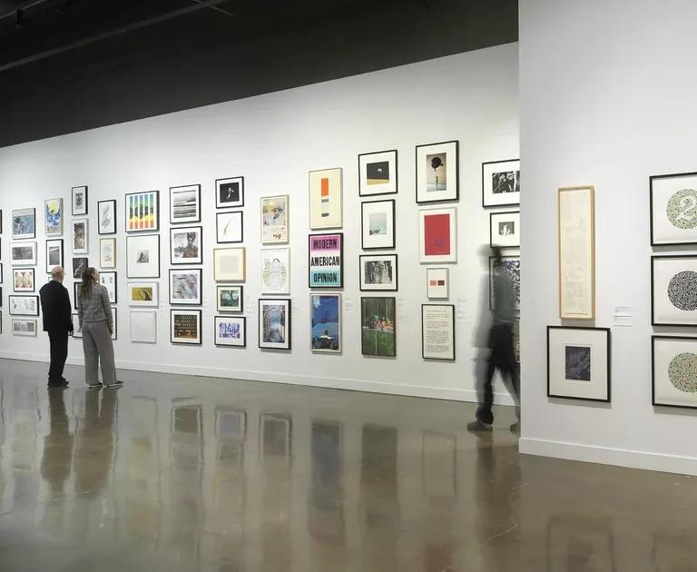 Installation view with framed artworks hung salon-style on the walls and three people looking at the art