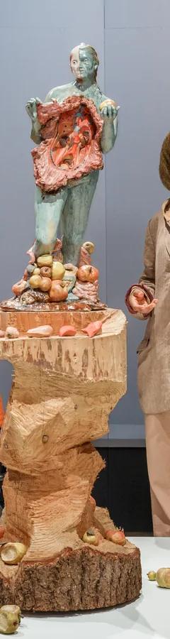 Two visitors look at sculptures of three women on large wooden pillars surrounded by apples; the central figure's body has been opened up and her insides are visible.