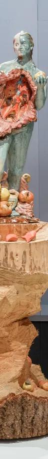 Two visitors look at sculptures of three women on large wooden pillars surrounded by apples; the central figure's body has been opened up and her insides are visible.
