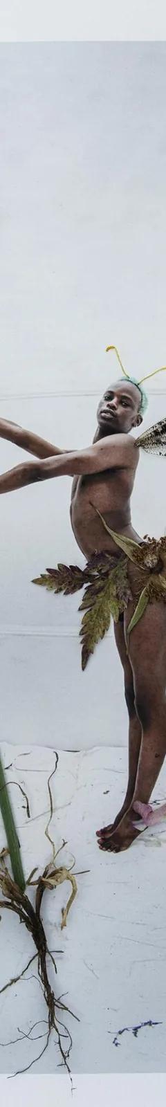 Colour photo of a black man dressed as a fairy holding the stalk of a plant with blue flowers, blue flowers at his feet and a bee to one side.