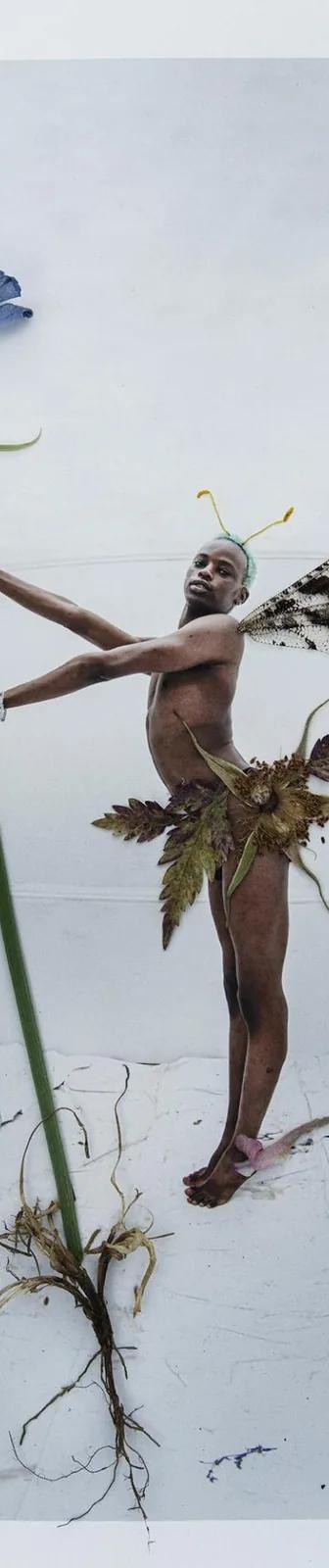 Colour photo of a black man dressed as a fairy holding the stalk of a plant with blue flowers, blue flowers at his feet and a bee to one side.