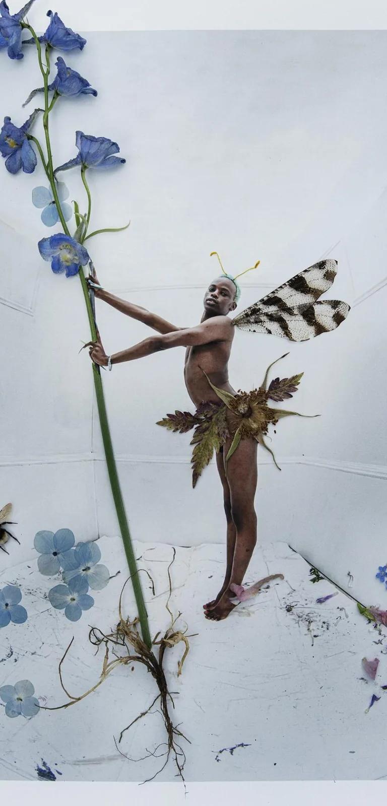 Colour photo of a black man dressed as a fairy holding the stalk of a plant with blue flowers, blue flowers at his feet and a bee to one side.