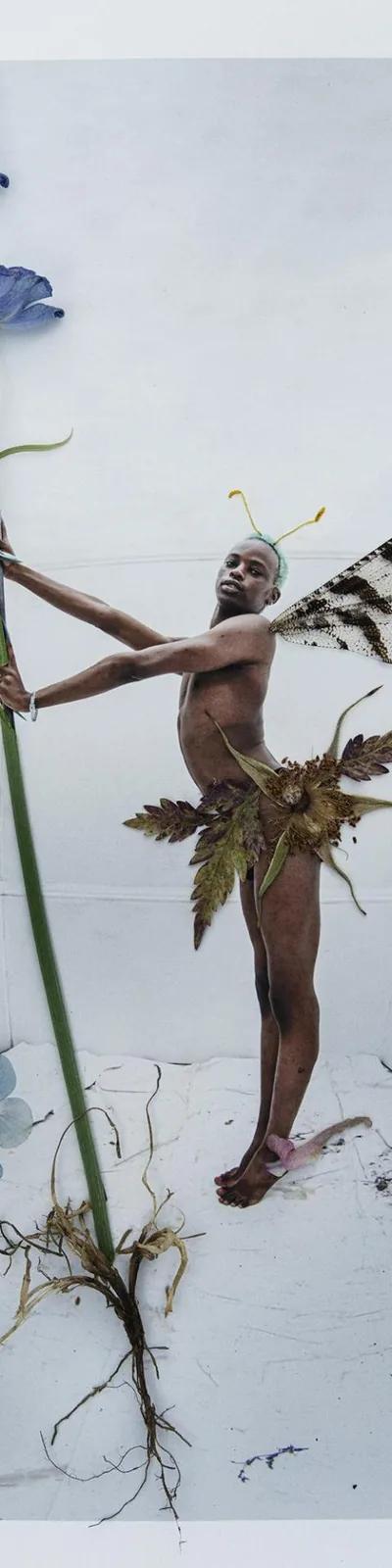 Colour photo of a black man dressed as a fairy holding the stalk of a plant with blue flowers, blue flowers at his feet and a bee to one side.