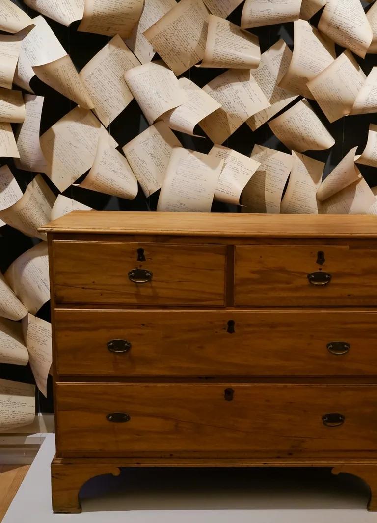 Pessoa's wooden chest of drawers in front of a wall of suspended handwritten manuscript pages