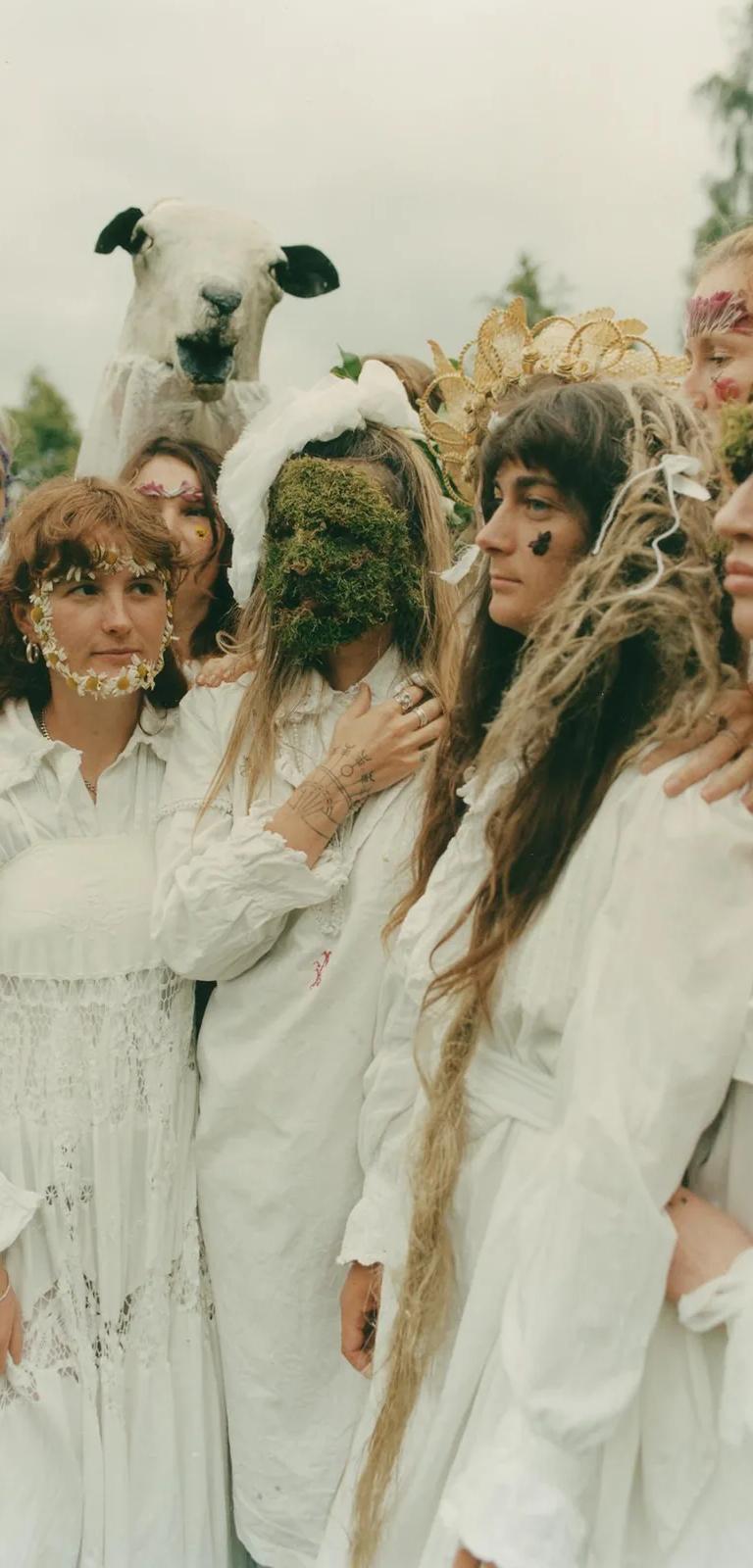 Colour photo of a group of young women outdoors in long white dresses, faces decorated with flowers and moss, a model sheep's head held up behind them.
